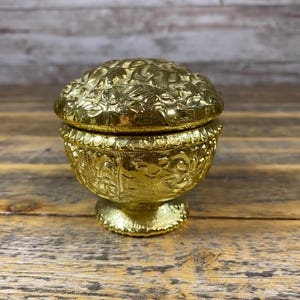 May include: A small, round, gold-colored metal trinket box with a lid. The exterior of the bowl and the lid feature an intricate, embossed floral and leaf pattern. The container rests on a circular pedestal base with a textured, scalloped edge.
