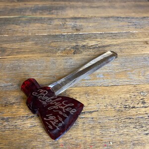 May include: A handcrafted, miniature axe with a clear handle and a dark red axe head. The axe head is etched with the name "Bobby Lee" and the year "1929". The axe is displayed on a rustic wooden surface.