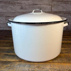 May include: A white enamel pot with a matching lid. The pot has a black trim around the rim and handles. The lid has a small handle. The pot is sitting on a wooden surface.