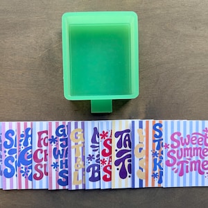 May include: A green rectangular soap mold is positioned above a row of colorful, striped cards. The cards feature the words "Sweet Summer Time" and other text in a retro font, with floral accents. The overall theme is summer.