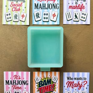 May include: A light mint green rectangular silicone mold positioned centrally, surrounded by six rectangular cards featuring striped backgrounds, Mahjong tile illustrations, and text including Mahjong Social Club, Let's play MAHJONG, Mahjong is mahlife, It's MAHJONG Time, BAM TIME, and Who wants to Mahj?