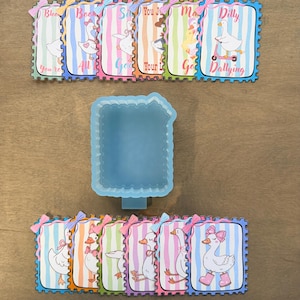 May include: A collection of colorful, whimsical goose-themed art pieces with text. The pieces are arranged around a blue, rectangular mold. Each piece features a goose illustration and a unique phrase, framed with a decorative border and bow.