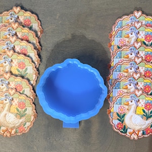 May include: A collection of decorative coasters featuring a charming goose design with floral and patchwork patterns. The coasters are arranged in stacks, alongside a blue, cloud-shaped mold. The coasters are approximately 4 inches in diameter.