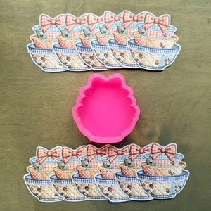 May include: A collection of stickers featuring decorative bowls with bows, in shades of blue, peach, and cream. A bright pink silicone mold in a scalloped shape is also present. The stickers and mold are arranged on a wooden surface.