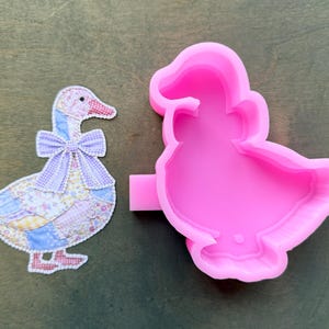 May include: A colorful goose-shaped applique next to a bright pink silicone mold. The applique features a patchwork design in pastel colors with a lavender bow. The mold is a detailed, reverse image of the applique.