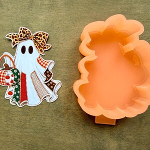 May include: A ghost-shaped cookie cutter in light orange plastic alongside a flat ghost-shaped cutout featuring a leopard print bow, a pumpkin-patterned basket, and a patchwork body with red, green, white, and brown fabric-style patterns.