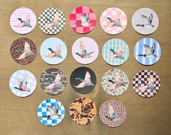 May include: A collection of seventeen circular stickers, each featuring a mallard duck in flight. The backgrounds vary, including patterns such as stripes, checkered squares, leopard print, camouflage, and solid colors in shades of blue, pink, and black.