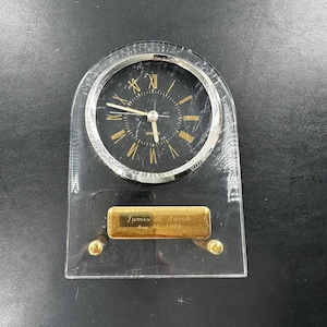 Peut inclure: Une horloge à quartz en acrylique transparent avec un cadran noir et des chiffres romains dorés. L'horloge a une lunette argentée et des accents dorés. Une plaque dorée à la base porte l'inscription "James & Sarah May 22 1999."