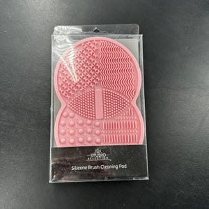 Puede incluir: Una almohadilla de limpieza de brochas de silicona rosa en un paquete de plástico transparente. La almohadilla tiene varias superficies texturizadas para limpiar brochas de maquillaje. El texto "STUDIO SELECTION" y "Silicone Brush Cleaning Pad" está incluido.