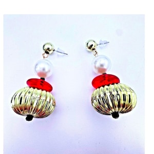 May include: A pair of gold-colored earrings with a unique design. Each earring features a gold ball stud, a white pearl, a red disc, and a large, ribbed, gold-colored, pumpkin-shaped bead. The earrings have a black bead at the bottom.
