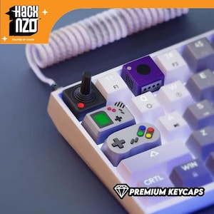 May include: A white mechanical keyboard featuring custom artisan keycaps shaped like a black joystick, a purple game console, a grey handheld gaming device, and a grey game controller. A white coiled cable is connected to the top of the keyboard.