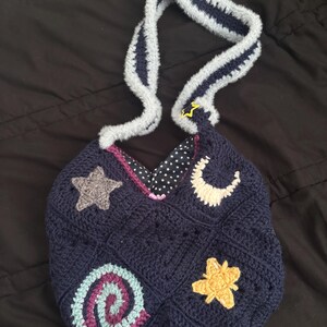 K&ouml;nnte beinhalten: Eine marineblaue H&auml;keltasche mit einem grau-blau gestreiften Henkel. Die Tasche hat gestickte Himmelsmotive, darunter ein grauer Stern, ein gelber Stern, ein wei&szlig;er Halbmond und eine Spirale. Das Innenfutter ist mit wei&szlig;en Sternen auf schwarzem Hintergrund gemustert.
