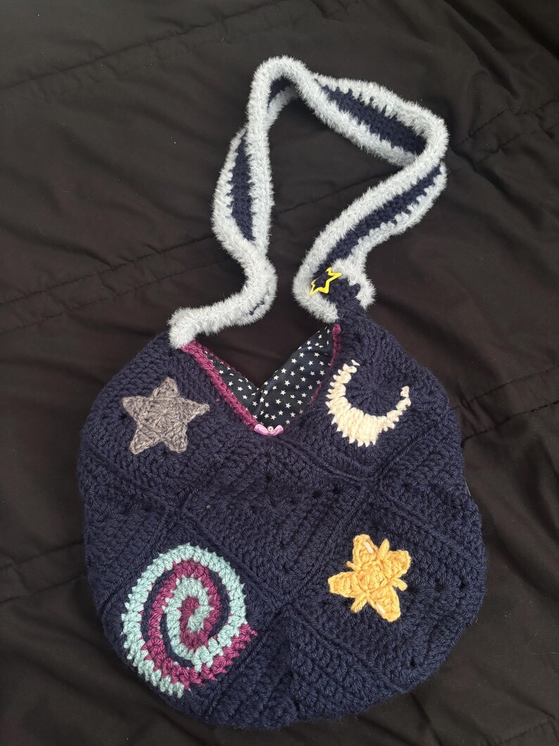K&ouml;nnte beinhalten: Eine marineblaue H&auml;keltasche mit einem grau-blau gestreiften Henkel. Die Tasche hat gestickte Himmelsmotive, darunter ein grauer Stern, ein gelber Stern, ein wei&szlig;er Halbmond und eine Spirale. Das Innenfutter ist mit wei&szlig;en Sternen auf schwarzem Hintergrund gemustert.