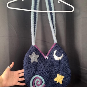 K&ouml;nnte beinhalten: Eine handgefertigte marineblaue H&auml;keltasche mit einem Himmelsmotiv. Die Tasche hat ein Stern-, Mond- und Spiraldesign. Die Tasche hat einen hellblauen und marineblauen Riemen und h&auml;ngt an einem wei&szlig;en Kleiderb&uuml;gel.