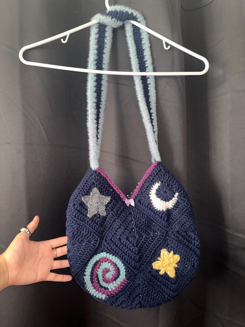 K&ouml;nnte beinhalten: Eine handgefertigte marineblaue H&auml;keltasche mit einem Himmelsmotiv. Die Tasche hat ein Stern-, Mond- und Spiraldesign. Die Tasche hat einen hellblauen und marineblauen Riemen und h&auml;ngt an einem wei&szlig;en Kleiderb&uuml;gel.