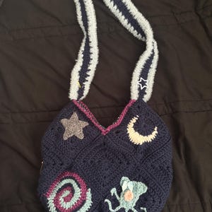 K&ouml;nnte beinhalten: Handgefertigte marineblaue H&auml;keltasche mit dekorativen Applikationen. Die Tasche hat einen Stern, einen Mond, eine Spirale und ein Seeungeheuer. Der Tr&auml;ger ist grau mit marineblauen Streifen. Ein einzigartiges Accessoire.