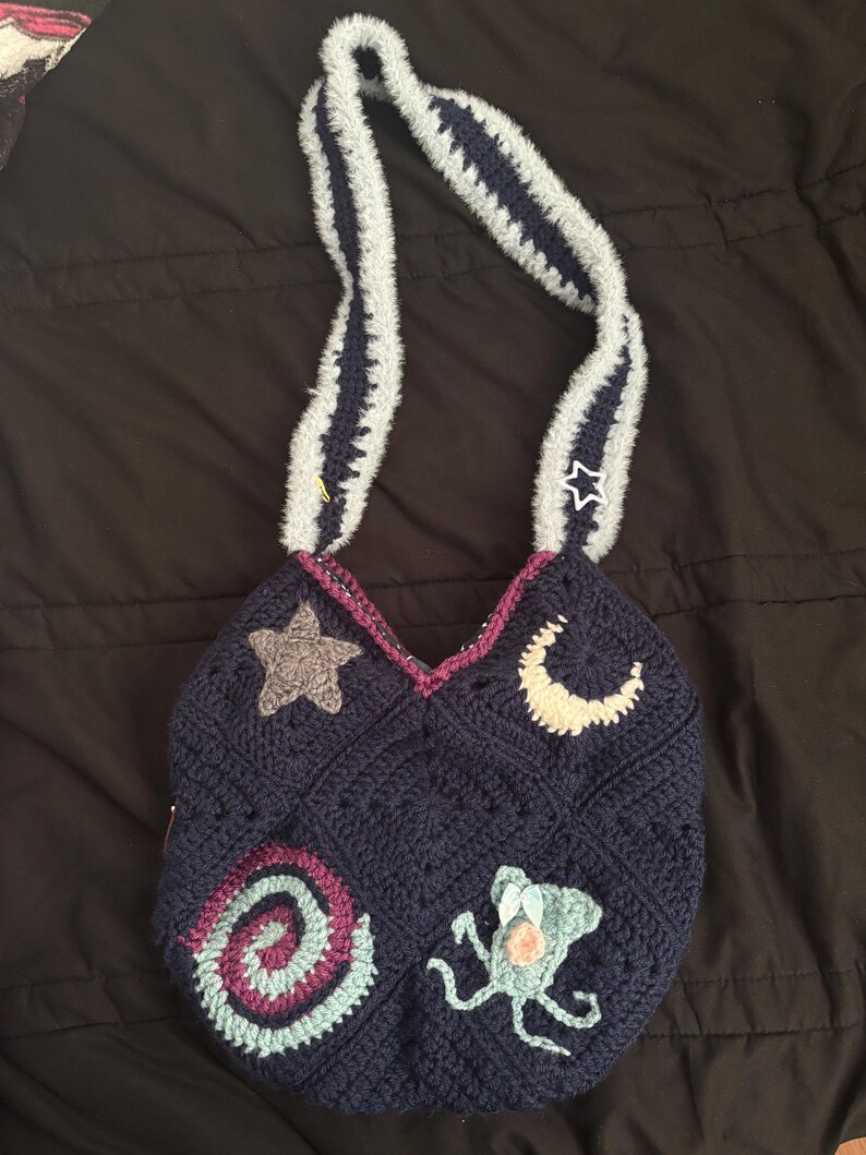 K&ouml;nnte beinhalten: Handgefertigte marineblaue H&auml;keltasche mit dekorativen Applikationen. Die Tasche hat einen Stern, einen Mond, eine Spirale und ein Seeungeheuer. Der Tr&auml;ger ist grau mit marineblauen Streifen. Ein einzigartiges Accessoire.