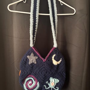 K&ouml;nnte beinhalten: Marineblaue H&auml;keltasche mit geometrischem Muster, verziert mit Stern-, Mond-, Spiral- und Meereskreaturen-Applikationen. Die Tasche hat hellblaue, flauschige Tr&auml;ger mit einem dunkelblauen Streifen. H&auml;ngt an einem wei&szlig;en Kleiderb&uuml;gel.