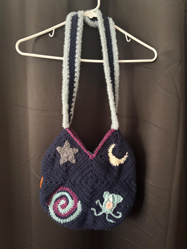 K&ouml;nnte beinhalten: Marineblaue H&auml;keltasche mit geometrischem Muster, verziert mit Stern-, Mond-, Spiral- und Meereskreaturen-Applikationen. Die Tasche hat hellblaue, flauschige Tr&auml;ger mit einem dunkelblauen Streifen. H&auml;ngt an einem wei&szlig;en Kleiderb&uuml;gel.