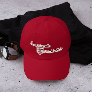 Könnte beinhalten: Rote Baseballkappe mit weiß gesticktem Text "Beechcraft Bonanza" und einem kleinen weißen gestickten Wappen mit einem Kreuz.