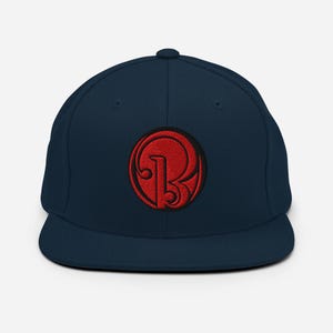 Könnte beinhalten: Marineblaue Baseballkappe mit flachem Schirm und einem roten, gestickten, kreisförmigen Logo. Das Logo zeigt einen stilisierten Buchstaben "B". Die Kappe hat eine strukturierte Passform und einen klassischen, lässigen Stil.