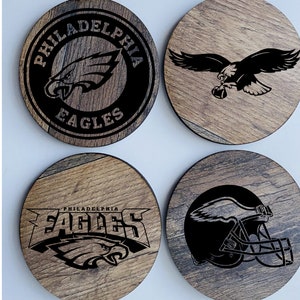 May include: Four round wooden coasters with black and white Philadelphia Eagles logos. The logos include a bird, a helmet, and the words "Philadelphia Eagles".