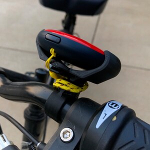 May include: A black bicycle handlebar with a red and black device attached to it. The device is secured with a black plastic mount and a yellow rope. The handlebar has a black grip with a white sticker that says 'E'. Bike & Scooter Mount | Secure Handlebar Clip for Outdoor Gaming and Events | 3 Size Options | Compatible with GO Plus + Device