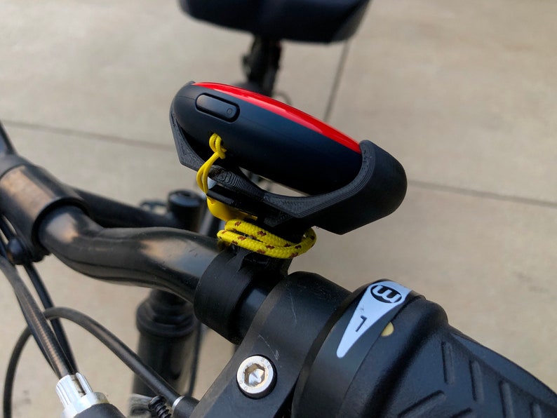 May include: A black bicycle handlebar with a red and black device attached to it. The device is secured with a black plastic mount and a yellow rope. The handlebar has a black grip with a white sticker that says 'E'. Bike & Scooter Mount | Secure Handlebar Clip for Outdoor Gaming and Events | 3 Size Options | Compatible with GO Plus + Device