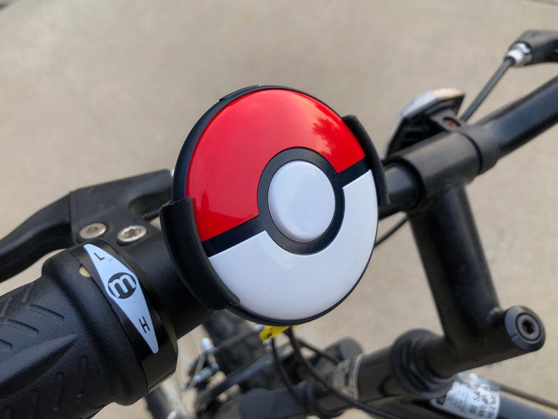 May include: A black bicycle handlebar with a red, black, and white Pokeball-shaped bell attached to it. The bell has a button on the side. The handlebar also has a black lever with a white 'L' and 'H' on it. Bike & Scooter Mount | Secure Handlebar Clip for Outdoor Gaming and Events | 3 Size Options | Compatible with GO Plus + Device