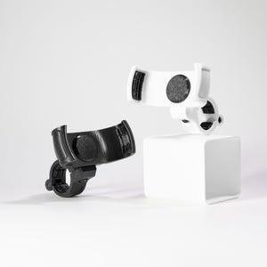 Two GO Plus + compatible handlebar mounts, rugged edition, shown in matte black and clean white. These 3D printed accessories are designed for bikes, scooters, strollers, and wheelchairs, featuring a secure snap-fit cradle and a circular handlebar clamp. The cradle includes interior padding for a non-slip grip on circular mobile gaming auto-catchers. Shown in a studio setting, these durable mounts provide a stable, hands-free solution for gaming during outdoor activities and travel.