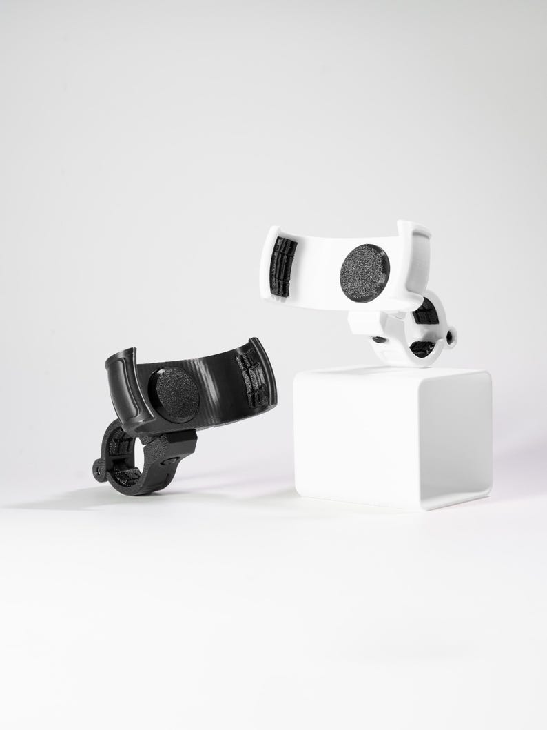 Two GO Plus + compatible handlebar mounts, rugged edition, shown in matte black and clean white. These 3D printed accessories are designed for bikes, scooters, strollers, and wheelchairs, featuring a secure snap-fit cradle and a circular handlebar clamp. The cradle includes interior padding for a non-slip grip on circular mobile gaming auto-catchers. Shown in a studio setting, these durable mounts provide a stable, hands-free solution for gaming during outdoor activities and travel.