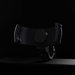 Dramatic dark lit product shot of a carbon fiber reinforced GO Plus + compatible handlebar mount against a black background. The matte-black mount features a carbon-infused texture and a curved snap-fit cradle with anti-slip padding. Designed for circular mobile gaming auto-catchers, the integrated bar-clamp at the base securely attaches to bikes, scooters, and strollers. This rugged accessory provides a stable, hands-free mounting solution for players during outdoor activities.
