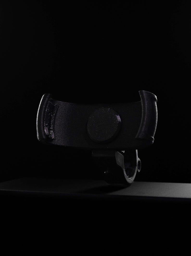 Dramatic dark lit product shot of a carbon fiber reinforced GO Plus + compatible handlebar mount against a black background. The matte-black mount features a carbon-infused texture and a curved snap-fit cradle with anti-slip padding. Designed for circular mobile gaming auto-catchers, the integrated bar-clamp at the base securely attaches to bikes, scooters, and strollers. This rugged accessory provides a stable, hands-free mounting solution for players during outdoor activities.