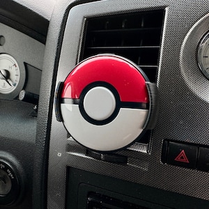 May include: A red, white, and black car air freshener shaped like a Pokeball. The air freshener is attached to the air vent in a car.