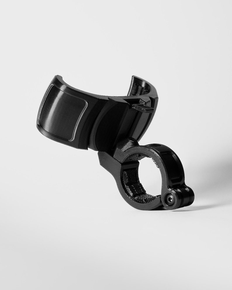 A professionally shot GO Plus + compatible handlebar mount, rugged edition, in gloss black. This angled product shot on a white background highlights the curved snap-fit cradle and the secure circular clamp at the base. The durable mounting accessory is designed to hold a circular mobile gaming auto-catcher on handlebars for bikes, scooters, and strollers, providing a stable, hands-free solution for active players.
