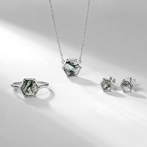 May include: A jewelry set featuring a ring, necklace, and earrings. Each piece has a hexagonal shape with a silver-colored metal frame. The gemstones have a green and white pattern. The necklace has a delicate chain, and the earrings are stud style.