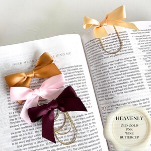 May include: A collection of satin ribbon bow bookmarks in old gold, pink, and wine colors, attached to gold-toned paper clips. The bookmarks are placed on open pages of a book with the word "Heavenly" on a white wax seal.