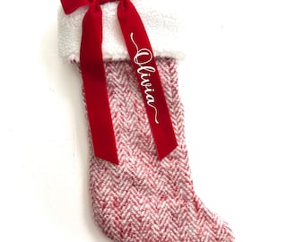 May include: A Christmas stocking with a red and white herringbone pattern. The top is trimmed with white faux fur. A red velvet ribbon is tied at the top with the name "Olivia" in white script. A brown leather loop is attached for hanging.