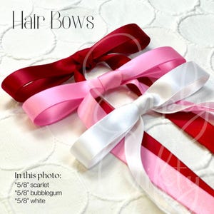 May include: Three hair bows in different colors: scarlet red, bubblegum pink, and white. Each bow is made of satin ribbon and tied in a classic bow shape. The bows are arranged on a white background.