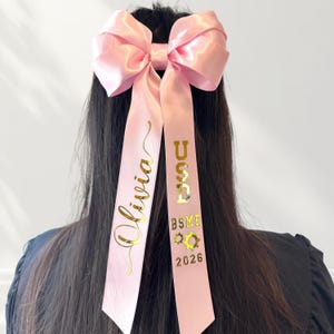 A close-up shot of a model wearing a pink convertible Grad Cap ribbon bow as a hair accessory. The bow is crafted from 1.5" double satin ribbon featuring reflective gold chrome personalization. One tail displays a name in elegant script, while the other tail features block lettering for the school name, field of specialty, and graduation year.