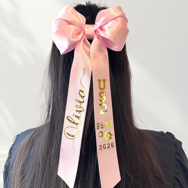 A close-up shot of a model wearing a pink convertible Grad Cap ribbon bow as a hair accessory. The bow is crafted from 1.5" double satin ribbon featuring reflective gold chrome personalization. One tail displays a name in elegant script, while the other tail features block lettering for the school name, field of specialty, and graduation year.