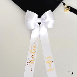A white personalized ribbon bow resting on a black mortarboard cap. The bow is crafted from double-satin white ribbon with gold chrome script lettering, featuring a name on one tail and the caduceus symbol with 'LPN' and graduation year on the other. The image is a flat-lay detail shot highlighting the metallic personalization and the detachable clip feature.