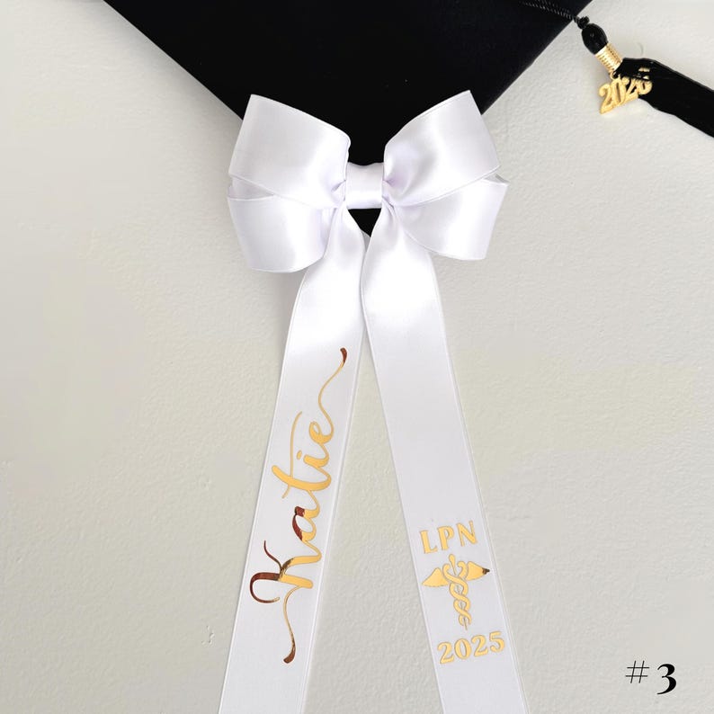 A white personalized ribbon bow resting on a black mortarboard cap. The bow is crafted from double-satin white ribbon with gold chrome script lettering, featuring a name on one tail and the caduceus symbol with 'LPN' and graduation year on the other. The image is a flat-lay detail shot highlighting the metallic personalization and the detachable clip feature.