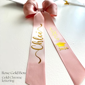 A rose gold personalized ribbon bow resting on a white mortarboard cap. The bow is crafted from double-satin ribbon with gold chrome script lettering, featuring a name on one tail and the graduation year on the other. The image is a flat-lay detail shot highlighting the metallic personalization and the detachable clip feature.
