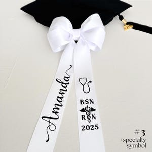 A white personalized ribbon bow resting on a black mortarboard cap. The bow is crafted from 1.5" double-satin ribbon with bold black lettering. The left tail features a name in elegant script, while the right tail is personalized with a stethoscope symbol, a caduceus with 'RN' and the graduation year. The image is a flat-lay detail shot highlighting the medical-themed personalization and the detachable clip.