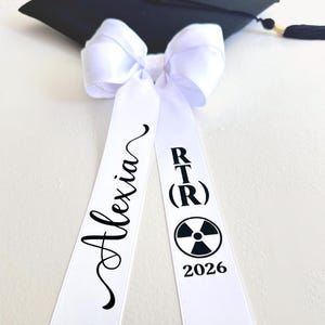 2026 Graduation Cap Bow personalized with name and field of study on the tails.  Large white coquette bow with the inscription in black vinyl, "alexia" and "RT(R) with the radiology symbol and date".  Laying flat with a black graduation cap with tassle.
