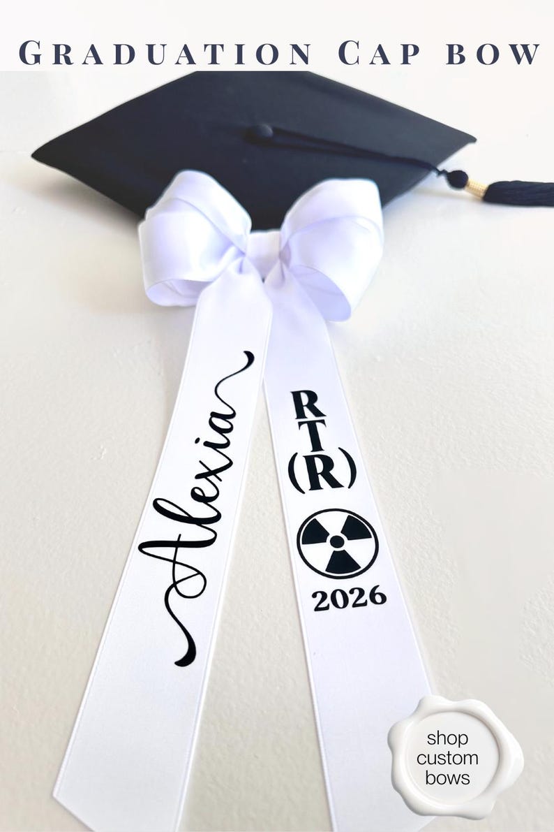 2026 Graduation Cap Bow personalized with name and field of study on the tails.  Large white coquette bow with the inscription in black vinyl, "alexia" and "RT(R) with the radiology symbol and date".  Laying flat with a black graduation cap with tassle.