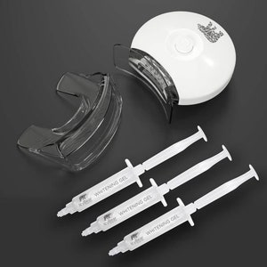 Puede incluir: Un kit de blanqueamiento dental con una luz LED blanca, un protector bucal transparente y tres jeringas de gel blanqueador. Las jeringas están etiquetadas como "Whitening Gel", la luz tiene un logotipo de oso.