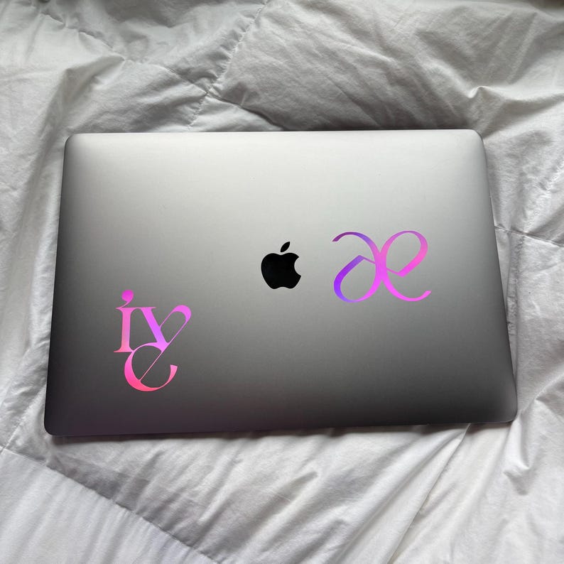May include: A silver laptop featuring a black Apple logo in the center. Two gradient-colored stickers are applied to the lid: one on the left displaying the text 'ive' in a stylized font, and one on the right showing the 'ae' ligature symbol.
