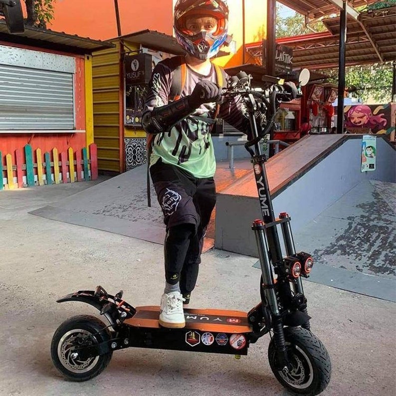 May include: A black electric scooter with orange accents. The scooter has two large wheels, a black handlebar with a red light, and a black deck with a YUME logo. The scooter is standing on a gray concrete surface.